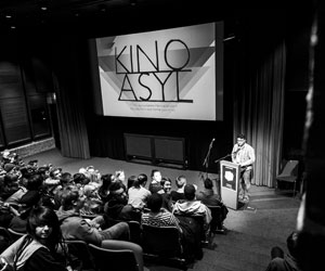 Zuschauer*innen sitzen vor einer Bühne. Auf der Bühne hängt ein Banner mit "Kino Asyl". Eine Person hält eine Rede an einem Rednerpult auf der Bühne.