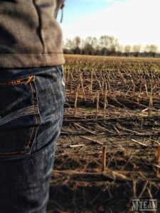 Linke Seite einer Person im Vordergrund, es sind ein Teil der Jeans und einer grauen Jacke zu erkennen, im Hintergrund ein abgeerntetes Feld. 